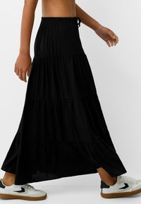 Schwarzer, gestufter Maxirock aus leichtem Stoff, mit einem elastischen Bund und einem Kordelzugverschluss. Kombiniert mit weißen und schwarzen Sneakers.