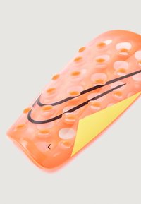 Espinillera de Nike en plástico naranja translúcido con puntos elevados, logotipo negro y un acento triangular amarillo. Tamaño marcado como 'L'.