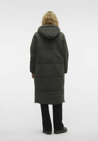 Abrigo largo estilo plumífero en verde oscuro, con capucha, textura acolchada y aberturas laterales, combinado con pantalones negros y botas negras gruesas.