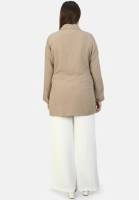 Beige long-sleeve jacket with a collar, made of textured fabric, paired with wide-leg white pants and square-heeled white shoes.