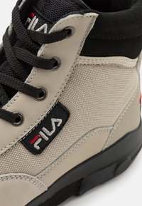 Sneaker high-top in tessuto beige con accenti neri, caratterizzata da una superficie testurizzata, lacci neri e una vistosa toppa con logo Fila.