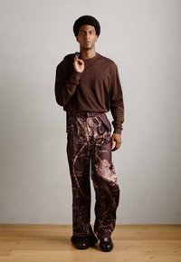 Camisa marrón de manga larga combinada con pantalones sueltos de corte amplio en marrón oscuro con patrones intrincados. Textura suave, con hardware mínimo.