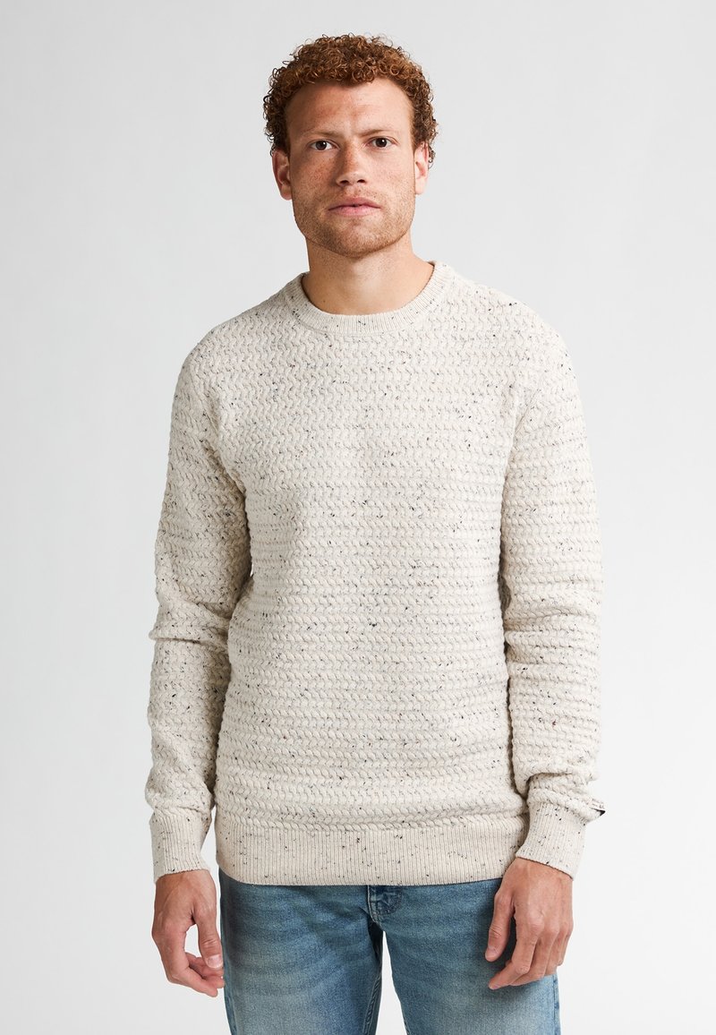 Crème kleurige gebreide trui met een structuurtje, lange mouwen en een ribbelboord bij de hals en mouwen. Draagt met blauwe denim jeans.