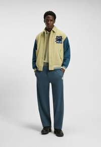 Jacket features a light green body with blue sleeves, embroidered logo on chest; paired with dark teal pants and black shoes.