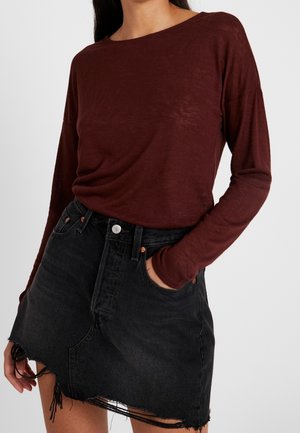 Kobieta ubrana w bordową bluzkę z długim rękawem, wpuszczoną w czarną, poszarpaną, asymetryczną spódnicę jeansową, z jedną ręką w kieszeni.