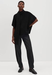 Donna in piedi con una mano sui fianchi, che indossa un maglione a collo alto nero con sciarpa integrata, pantaloni neri a gamba larga e mocassini neri.