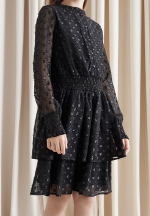 Frau trägt ein durchsichtiges schwarzes Langarmkleid mit strukturiertem Blumenmuster, geraffter Taille, gerafften Bündchen und mehrlagigem Rock vor beigem Vorhanghintergrund.