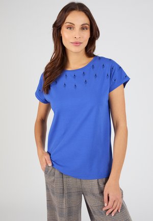 Mujer con camiseta de manga corta de color azul brillante con pequeños patrones de diamantes negros cerca del escote y pantalones de cuadros, de pie con una mano en el bolsillo.