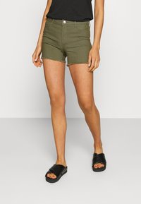 Green denim shorts with frayed hem, fitted design, one button closure, and front pockets, paired with black slide sandals.