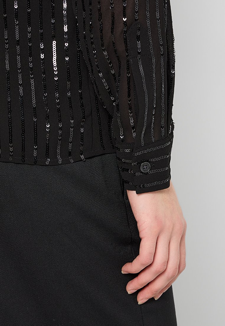 Zwarte sheer blouse met verticale rijen pailletten, voorzien van een knoopdetail bij de manchetten en een gladde textuur, gecombineerd met een donkere stof eronder.