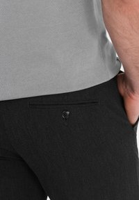 Grijze fitted shirt gecombineerd met zwarte textuurbroek met een achterzak met knoopsluiting, wat de minimalistische stijl benadrukt.
