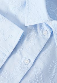 Camisa de botones azul claro con patrones florales bordados, que presenta botones blancos y una textura de algodón suave.
