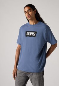 Mėlyna medvilninė marškinė su trumpomis rankovėmis ir apvaliu kaklu. Priekyje yra juodas Levi's logotipas. Dėvima su pilkais džinsais.