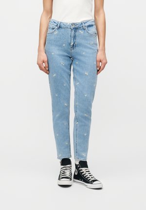 Kvinne iført lyseblå jeans med små broderte prestekrager, hvit topp og svarte høye sneakers, stående mot ensfarget bakgrunn.