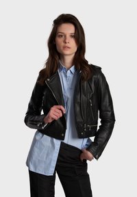 Black leather cropped jacket with zippered pockets, silver hardware, and a lapel collar, paired with a light blue button-up shirt.