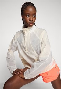 Veste de pluie blanche légère et semi-transparente avec un col haut et fermeture éclair. Équipée de poignets élastiques et d'un logo discret. Assortie à un short orange fluo.