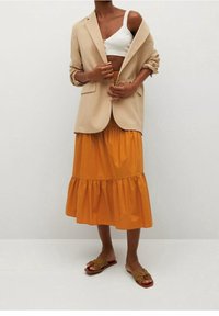 Blazer beige sopra un crop top bianco, abbinato a una gonna arancione a balze e sandali verde oliva. Tessuto liscio con texture a strati.
