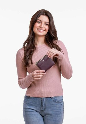 Maroon leather wallet with a green zipper, held by a person in a light pink ribbed sweater against a plain white background.