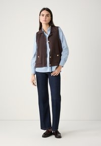 Braune ärmellose Weste mit goldenen Knöpfen, über einem hellblauen und weißen gestreiften Hemd, kombiniert mit dunkelblauen Straight-Leg-Jeans.