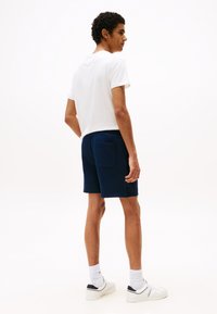 Marineblaue Shorts mit einer Gesäßtasche, kombiniert mit einem schlichten weißen T-Shirt. Das Modell steht mit dem Rücken zur Kamera vor einem einfarbigen Hintergrund und trägt weiße Sportchuhe.