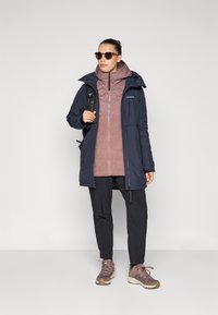 Woman wearing sunglasses, navy blue jacket, mauve quilted and fleece jacket, black pants, mauve hiking shoes, and carrying a black backpack.