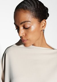 Top in tessuto beige drappeggiato con una consistenza morbida, caratterizzato da un ampio scollo. Il soggetto presenta una pelle liscia e indossa un piccolo orecchino scuro.