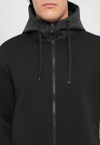Svart zip-up hettegenser med en kviltet svart hette, snøring og frontglidelås. Stoffet er glatt med en matt overflate.