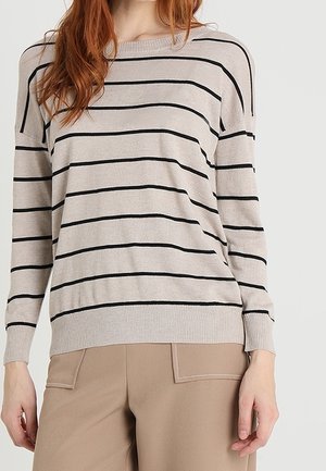 Frau trägt einen beigen Pullover mit schwarzen horizontalen Streifen und hellbraune Hose mit Vordertaschen, steht vor neutralem Hintergrund.