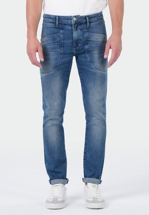 Blue denim jeans with curved front pockets, rolled cuffs, worn with white sneakers and a white shirt, shown on a standing person.