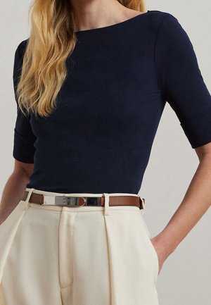 Vrouw die een marineblauw shirt met korte mouwen draagt, ingestopt in een crème kleurige broek met hoge taille en een dunne bruine riem.