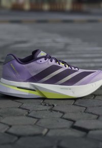 Μωβ και κίτρινο παπούτσι για τρέξιμο Adidas σε πλακόστρωτο δρόμο, δείχνοντας πλευρική όψη με πλέγμα στο πάνω μέρος και λεπτομέρειες μαλακής σόλας.