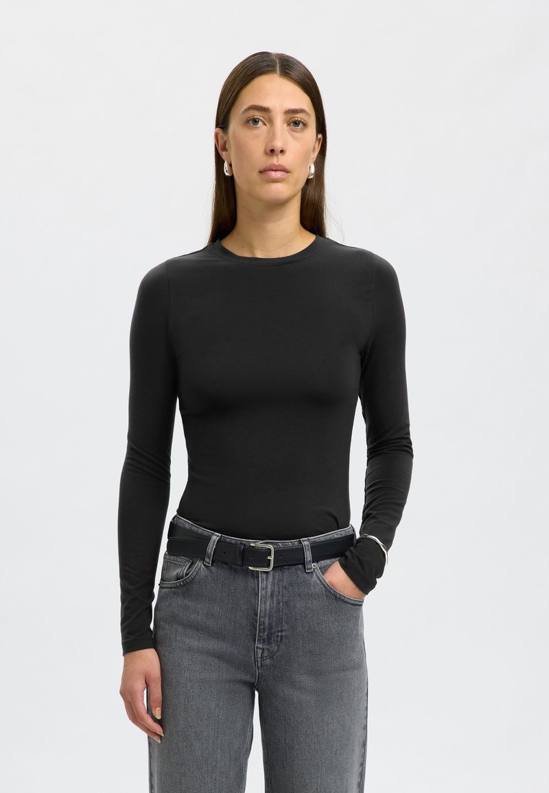 Juodas ilgarankovis prigludęs megztinis iš minkštos medžiagos, derinamas su aukštu liemeniu pilkais džinsais ir juodu diržu su sidabriniu sagtimi.