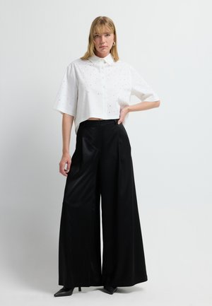 Witte kort geknipte blouse met subtiele versieringen, gecombineerd met hoge zwarte satijnen wijde broek en zwarte puntige hakken.