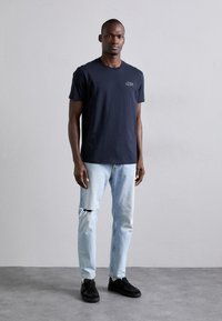 Camiseta de algodón azul marino con cuello redondo, combinada con jeans rasgados de un azul claro y zapatillas negras, de pie frente a un fondo plano.