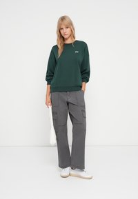 Sweatshirt verde escuro com logotipo "GAP" branco, combinado com calças cargo cinzentas com bolsos laterais e corte relaxado. Usado com sapatilhas brancas.