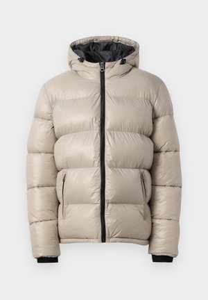 Personne portant une doudoune beige avec une fermeture éclair noire, un pantalon cargo beige et des baskets blanches, debout sur fond blanc.