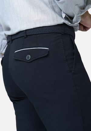 Man draagt marineblauwe broek met knoopsluiting aan achterzak en geweven riem, gecombineerd met lichtgrijs overhemd dat in de broek is gestopt.