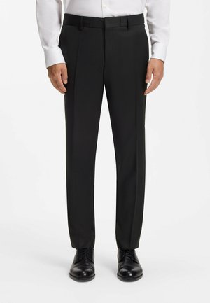 Homme portant un pantalon de costume noir, une chemise blanche boutonnée et des chaussures habillées noires, debout sur un fond blanc uni.