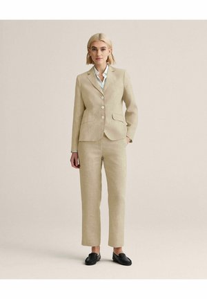 Kvinde stående iført en beige skræddersyet dragt med en hvid skjorte og sorte loafers, hænder afslappede, neutral baggrund.