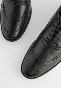 Los zapatos de vestir de cuero negro cuentan con un diseño de punta de ala, detalles decorativos de brogue, punteras redondas y un acabado liso con cierres de cordones.