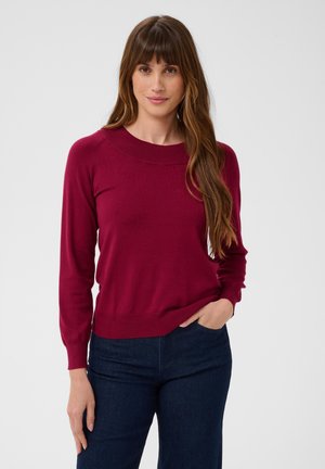 Femme aux longs cheveux bruns portant un pull bordeaux et un jean bleu foncé, debout devant un fond clair uni.