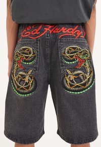 Black denim shorts feature vibrant snake embroidery on the back pockets, with "Ed Hardy" text stitched in red above.