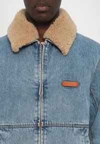 Denim jakk, millel on beež valejäägerkaelus, ees lukk ja pruun nahast logo silt. Kahvatud sinine värv, tekstuurne kangast detail.