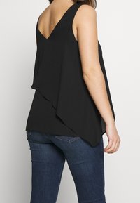 Svart ärmlös topp med V-formad bakdel, lagerdesign, flödande tyg och längre bakre fåll, kombinerad med mörka jeans.