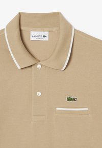 Beige bomullspiké med vit kragekant, prydd med en grön broderad krokodil logo på vänster bröst och två knappar.