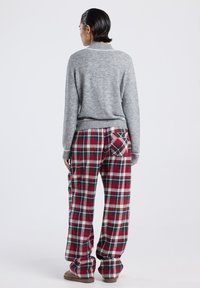 Personne portant un pull gris et un pantalon à carreaux rouges, verts et blancs avec une poche arrière, se tenant devant un fond léger uni.