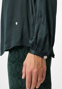 Mørkegrøn silke bluse med blød tekstur, med puffede ærmer og en juvelbesat knapdetalje ved manchetten for ekstra pynt.