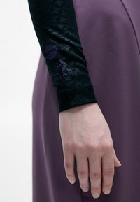 Melna blūze ar garām piedurknēm un faktūru, rotāta ar purpura akcentiem, saskaņota ar vienkrāsainu purpura kleitu. Piedurknē ir redzama zīmola apzīmējums.