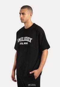Camiseta de algodón negra con mangas cortas, que presenta el texto blanco "SMILODOX ATHL. WEAR" en el pecho. Ajuste casual, diseño sencillo.