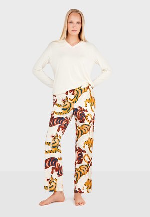 Femme aux longs cheveux blonds portant un haut blanc à manches longues et un pantalon à motifs avec des imprimés tigres multicolores, debout pieds nus.